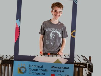 A boy with short red hair and wearing a grey t-shirt with a white print plays a glockenspiel. He stands in between a customised National Open Youth Orchestra Instagram frame.